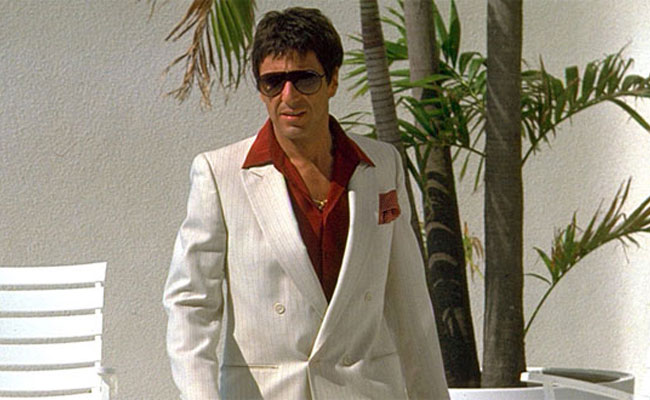 Man in a white suit, sunglasses, and red shirt, outdoors.